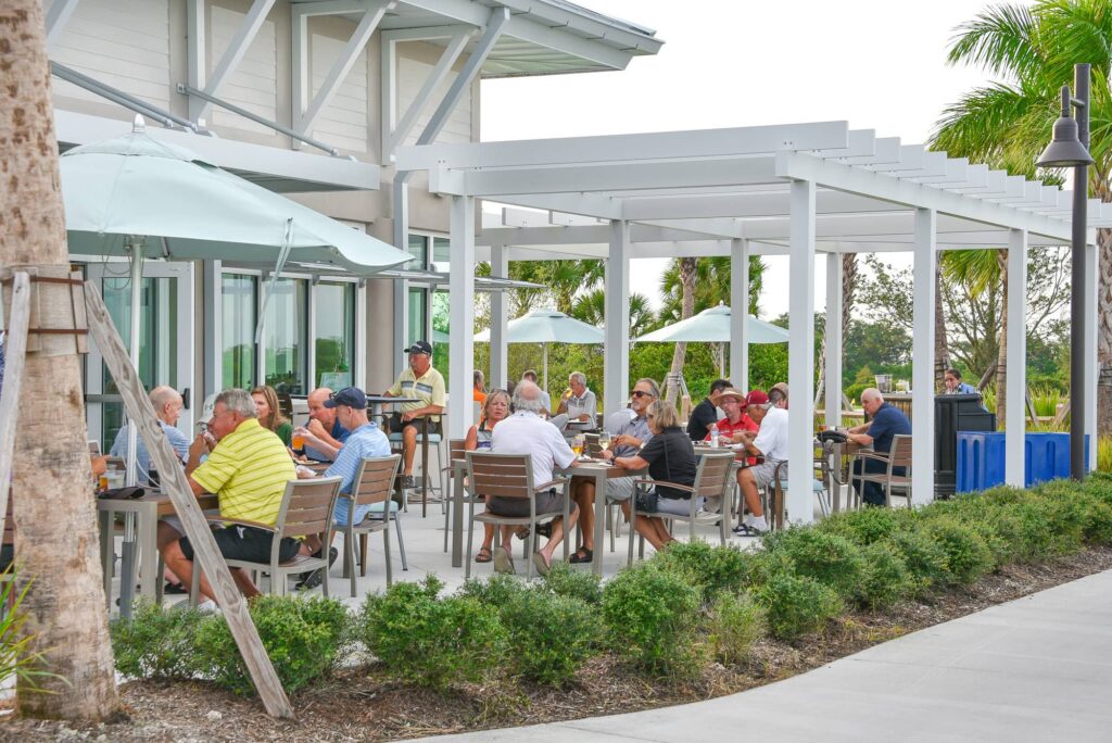 An outdoor dining area with a concrete floor and a large white wooden pergola. Multiple groups of people are seated at square wooden tables with modern grey chairs. Several light blue patio umbrellas provide extra shade. The building has large glass doors, and the perimeter is lined with green bushes and tall palm trees under an overcast sky.