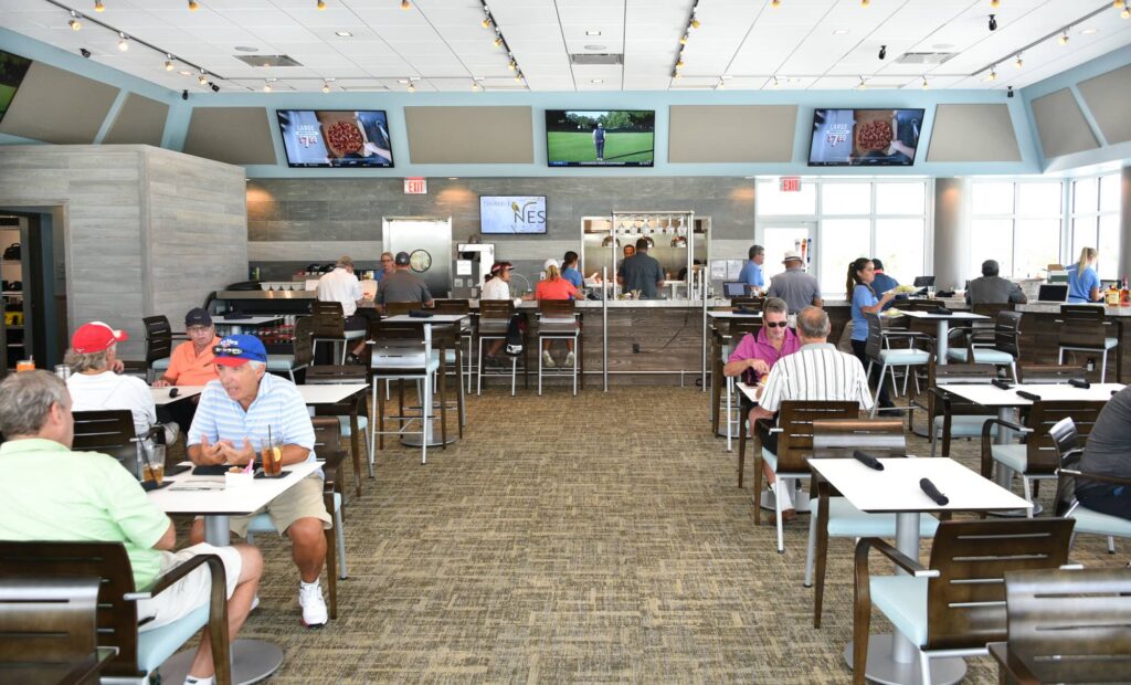 Interior view of a spacious, contemporary restaurant with a neutral color palette. Groups of people, mostly men in casual golf attire, are seated at square white tables and a long bar area. The background features a gray wood-paneled wall with four large flat-screen TVs showing various broadcasts. High ceilings with track lighting and large windows on the right create a bright, airy atmosphere. The flooring is a textured tan carpet, and the seating consists of modern dark wood chairs with light blue cushions.